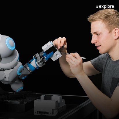 Ein junger Mann in grauem T-Shirt arbeitet konzentriert an einem weißen Kollaborationsroboter mit blauen Akzenten in einer technischen Arbeitsumgebung. Im Hintergrund sind Monitore mit technischen Spezifikationen sichtbar, während er präzise Einstellungen am Roboterarm vornimmt.