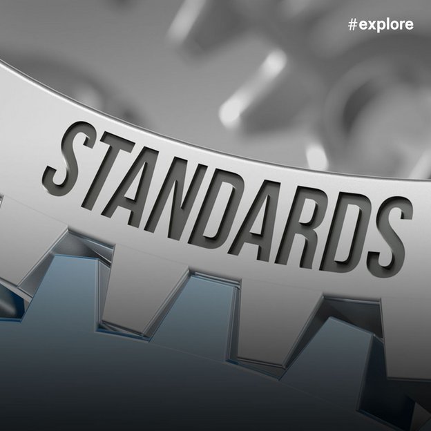 Ein metallisches Zahnrad mit dem eingraviertem Wort "STANDARDS" im Vordergrund, mit weiteren unscharfen Zahnrädern im Hintergrund in Grau- und Blautönen.