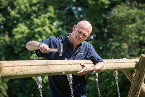 Ein Mann in dunkler Arbeitskleidung hämmert mit einem Hammer an einem Holzbalken. Ketten hängen von der Holzkonstruktion herab, die Teil eines Spielgeräts zu sein scheint.