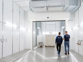 Zwei Techniker in TÜV NORD-Arbeitskleidung gehen durch eine moderne weiße Industriehalle mit technischen Geräten.
