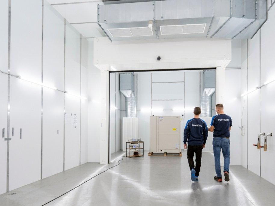 Zwei Techniker in TÜV NORD-Arbeitskleidung gehen durch eine moderne weiße Industriehalle mit technischen Geräten.