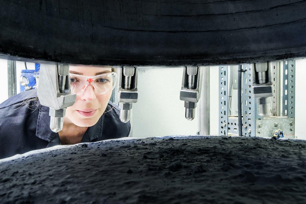 Wissenschaftlerin mit Schutzbrille betrachtet Materialprobe unter industrieller Prüfmaschine in einem Forschungslabor.