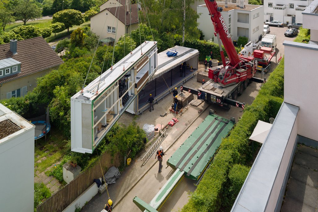 Luftaufnahme einer Baustelle, auf der modulare Wohneinheiten mit einem Kran installiert werden. Mehrere Arbeiter und vorgefertigte Gebäudeteile sind zu sehen.
