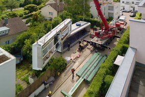 Luftaufnahme einer Baustelle, auf der modulare Wohneinheiten mit einem Kran installiert werden. Mehrere Arbeiter und vorgefertigte Gebäudeteile sind zu sehen.