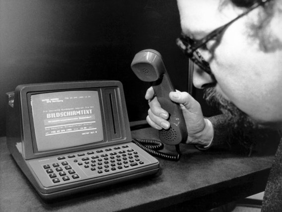Schwarz-weiß Foto eines Mannes mit Brille, der an einem frühen Computer mit Bildschirm und Tastatur arbeitet. Er hält einen kabelgebundenen Telefonhörer in der Hand.