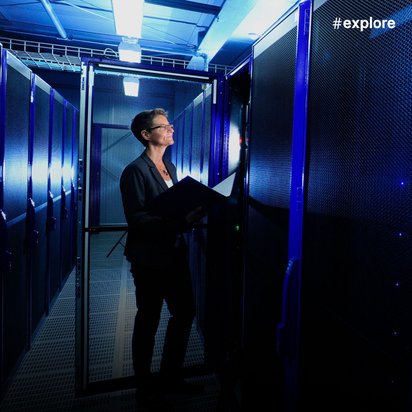 Eine Frau mit Brille und Laptop steht in einem modernen Rechenzentrum zwischen hohen Serverracks mit blauer LED-Beleuchtung. Die professionelle IT-Umgebung zeigt die technische Infrastruktur für Datenverarbeitung und Cloud-Services.