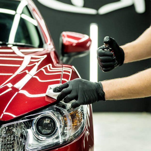 Ein rotes Auto wird von einer Person mit schwarzen Handschuhen sorgfältig poliert. Die glänzende Oberfläche des Fahrzeugs reflektiert das Licht, während die Person ein Pflegemittel aufträgt, um den Lack zu schützen und zu verschönern.