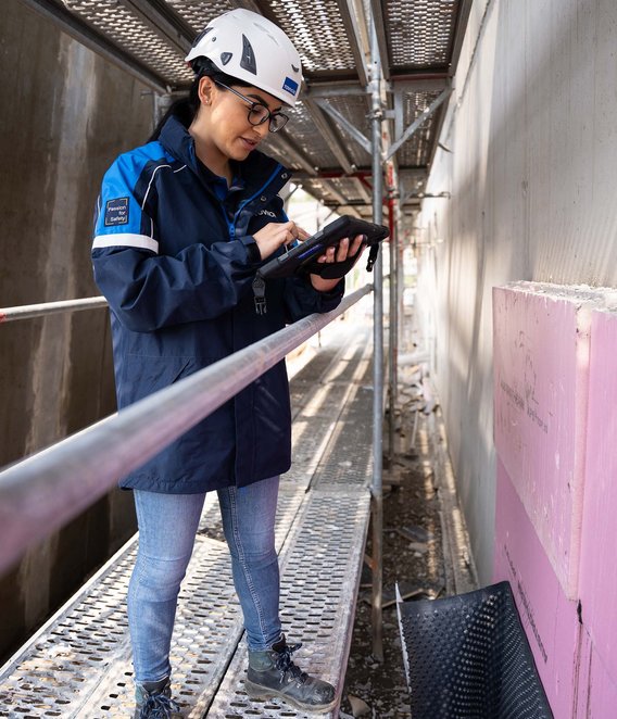 TÜV NORD-Mitarbeiterin mit Helm prüft bauliche Details auf einer Baustelle und dokumentiert mögliche Baumängel.