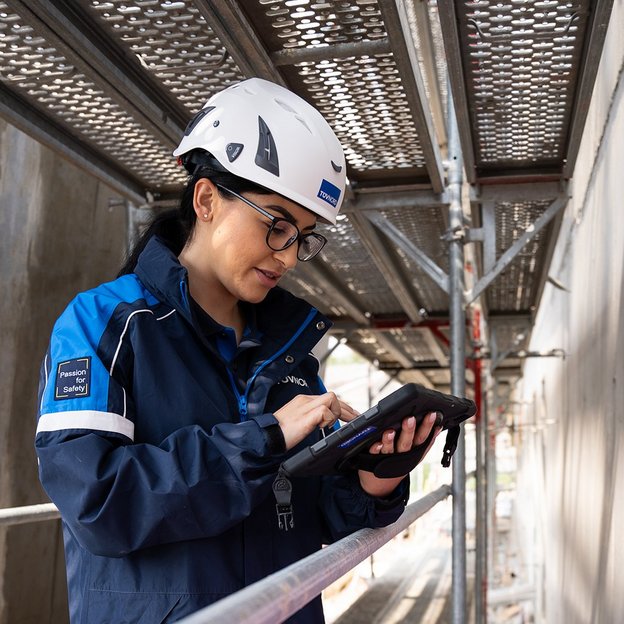 TÜV NORD-Mitarbeiterin mit Helm prüft bauliche Details auf einer Baustelle und dokumentiert mögliche Baumängel.