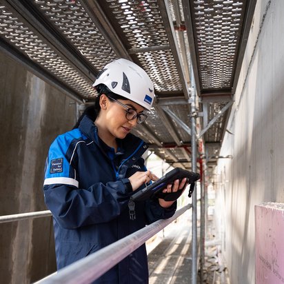 TÜV NORD-Mitarbeiterin mit Helm prüft bauliche Details auf einer Baustelle und dokumentiert mögliche Baumängel.