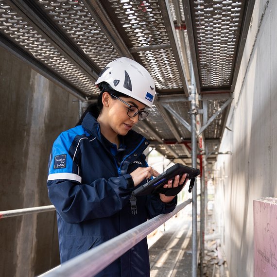 TÜV NORD-Mitarbeiterin mit Helm prüft bauliche Details auf einer Baustelle und dokumentiert mögliche Baumängel.