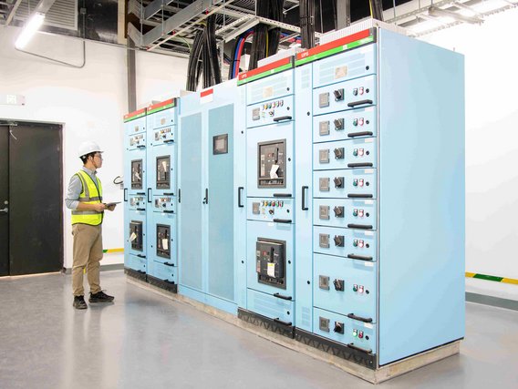 Techniker im Sicherheitshelm und Warnweste überprüft elektrische Schalttafeln in einem Kontrollraum.