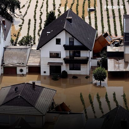 Luftaufnahme einer Hochwassersituation mit mehreren Häusern, die von braunem Flutwasser umgeben sind. Die Gebäude mit dunklen Dächern stehen teilweise im Wasser, während im Hintergrund Weinbergreihen zu sehen sind.