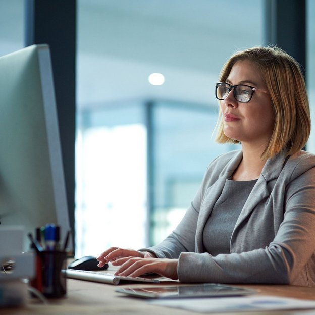 Frau mit Brille arbeitet konzentriert an einem Desktop-Computer in einem modernen Büro.