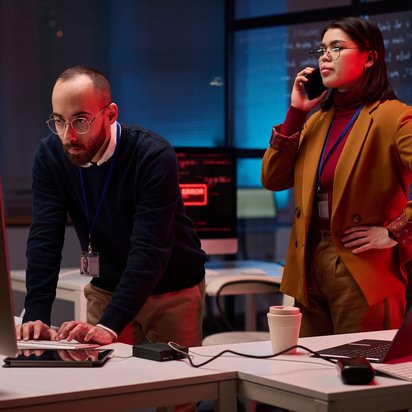 Zwei IT-Fachkräfte analysieren Sicherheitsvorfälle an Monitoren in einem abgedunkelten Büro.