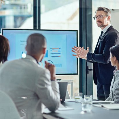 Geschäftsmann präsentiert vor Kollegen Diagramme auf einem großen Bildschirm im Konferenzraum.