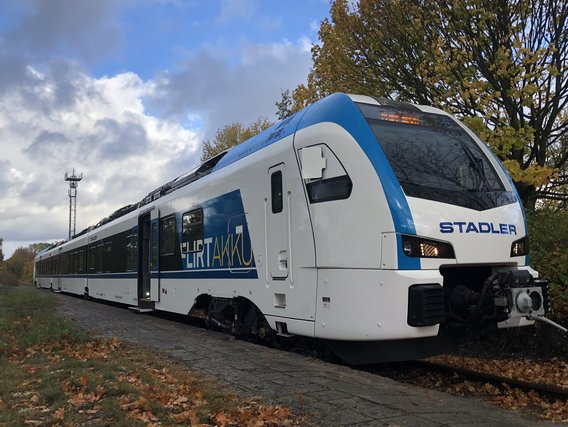 Moderner weiß-blauer Regionalzug der Marke Stadler mit Schriftzug "LHRTAVMU" steht auf Gleisen vor herbstlichen Bäumen und bewölktem Himmel.