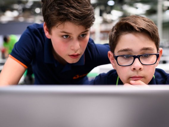 Zwei Jungen schauen auf einen Computerbildschirm. Ein Junge zeigt auf den Bildschirm, während der andere, der eine Brille trägt, zuhört. Sie programmieren vermutlich einen kleinen Roboter.