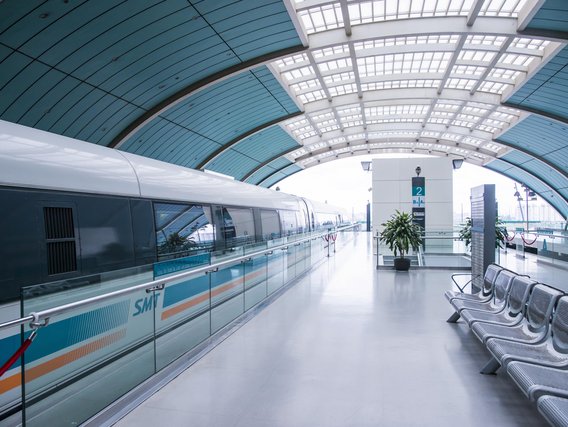 Moderne U-Bahn-Station mit türkisblauer Magnetbahn und gewölbter Glasdecke. Wartesitze aus Metall stehen rechts auf dem hellen Bahnsteig, Bahnsteigkante ist mit Glaswand gesichert.