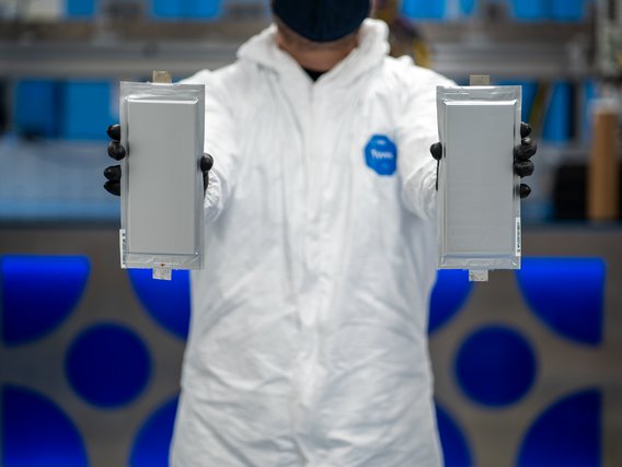 Eine Person in weißem Schutzanzug und blauer Maske hält zwei flache, rechteckige Batteriezellen oder Akkupacks in schwarzen Handschuhen vor einem Hintergrund mit blauen Markierungen.