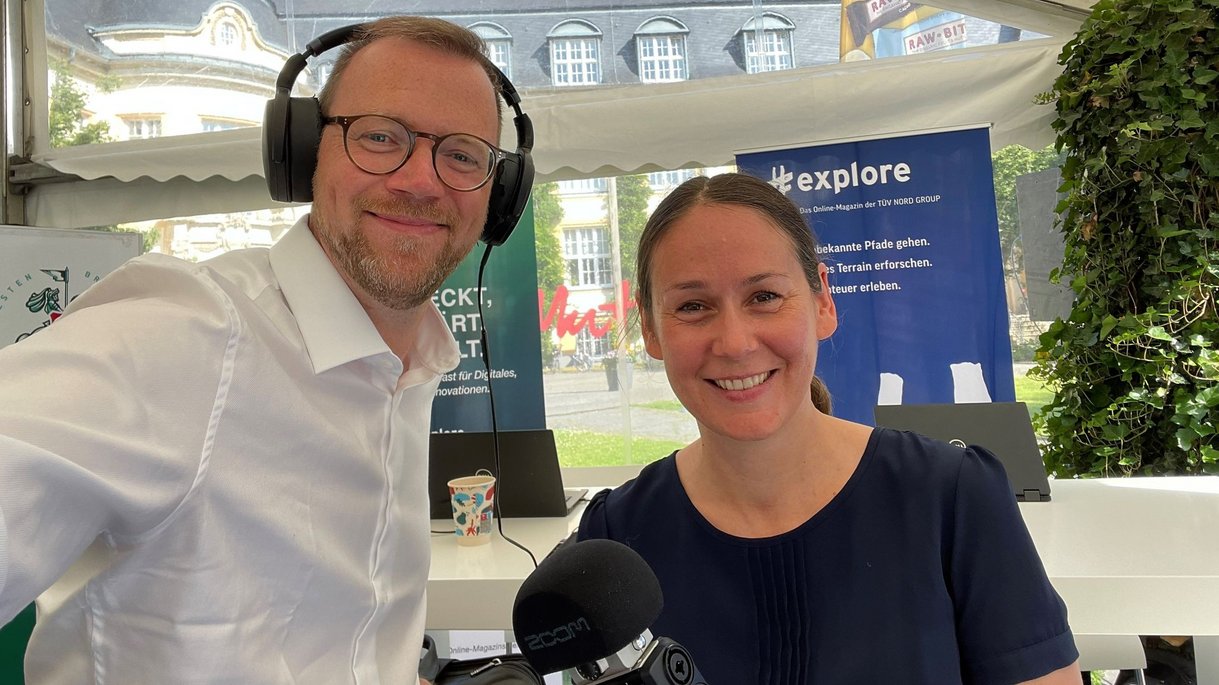 Carolin Roterberg und Martin Brüning für einen Podcast oder Interview, mit sichtbaren Bannern, darunter "explore" von TÜV NORD GROUP.