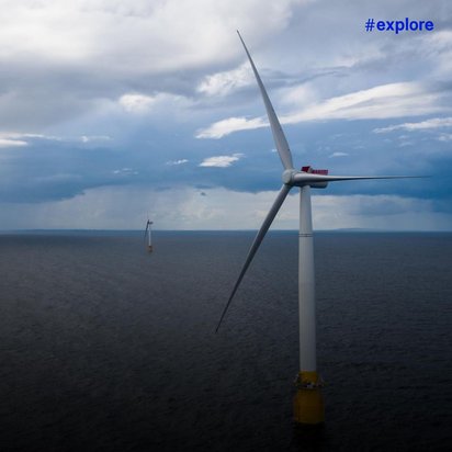 Aerial view of multiple offshore wind turbines spread across dark ocean waters under a dramatic cloudy sky. The turbines appear as white structures with yellow foundation bases extending to the horizon.