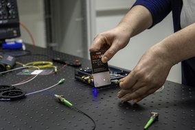 Eine Person hält eine Thorlabs Detektorkarte über einen Tisch mit technischen Geräten und Kabeln. Die Karte zeigt einen gelben Lichtpunkt, während die Person ein Kabel mit einem grünen Anschluss in der anderen Hand hält. Auf dem Tisch befinden sich weitere Kabel und elektronische Komponenten.