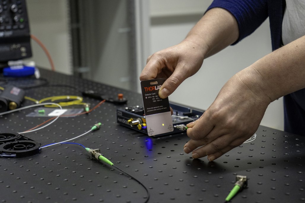 Eine Person hält eine Thorlabs Detektorkarte über einen Tisch mit technischen Geräten und Kabeln. Die Karte zeigt einen gelben Lichtpunkt, während die Person ein Kabel mit einem grünen Anschluss in der anderen Hand hält. Auf dem Tisch befinden sich weitere Kabel und elektronische Komponenten.