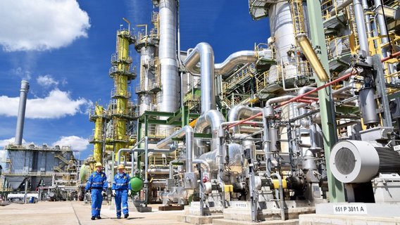 Zwei Männer in blauen Arbeitsanzügen und mit weißen Schutzhelmen laufen nebeneinander vor dem Hintergrund eines Chemieparks. Es sind diverse technische Anlagen unter blauen Himmel zu sehen.