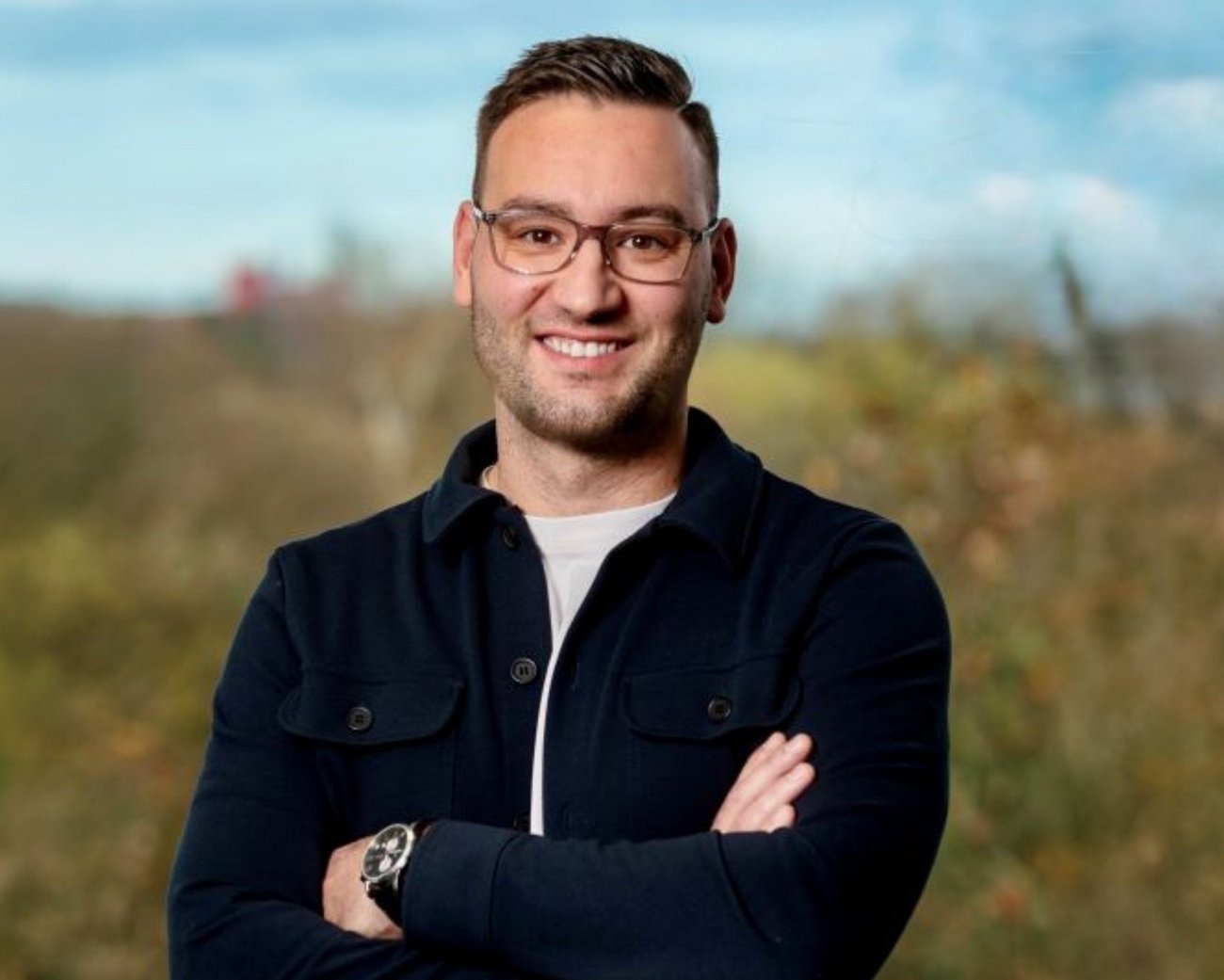 Eine Person steht mit verschränkten Armen im Freien und trägt eine dunkle Jacke über einem hellen T-Shirt. Der Hintergrund zeigt eine natürliche Landschaft mit Bäumen und einem bewölkten Himmel.