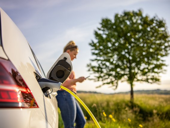 Ein Elektroauto wird mit einem gelben Ladekabel geladen, während eine Frau daneben auf ihr Smartphone schaut. Im Hintergrund steht ein grüner Baum in einer ländlichen Landschaft.
