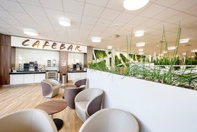 Moderne Cafeteria mit der Aufschrift "Cafebar" über einer Theke mit mehreren Kaffeeautomaten. Im Vordergrund sind stilvolle, runde Sitzgelegenheiten und Tische zu sehen. An der Wand sind grüne Pflanzen dekorativ angebracht.