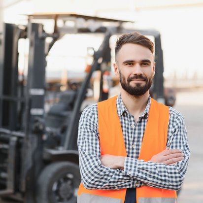 Lagerarbeiter in Sicherheitsweste steht vor einem Gabelstapler im Lagerbereich.