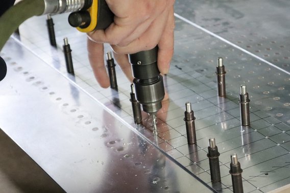Eine Person verwendet eine Bohrmaschine, um präzise Löcher in ein Metallstück zu bohren, das mit mehreren Halterungen versehen ist. Die Szene zeigt technische Fertigkeiten in der Luftfahrt.