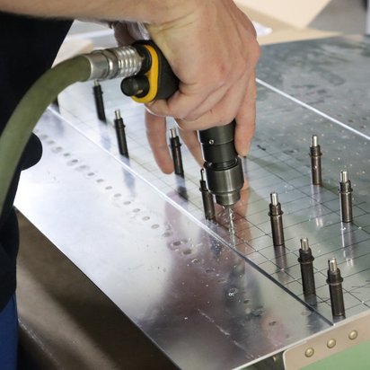 Eine Person verwendet eine Bohrmaschine, um präzise Löcher in ein Metallstück zu bohren, das mit mehreren Halterungen versehen ist. Die Szene zeigt technische Fertigkeiten in der Luftfahrt.