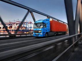 Ein blauer LKW mit rotem Container fährt über eine Stahlbrücke, im Hintergrund ist ein Hafen mit Kränen zu sehen.
