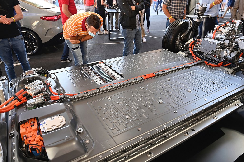 Eine große Elektrofahrzeug-Batterie wird auf einer Ausstellung präsentiert. Eine Person mit orangegestreiftem Shirt und Gesichtsmaske betrachtet den massiven Akku mit sichtbaren Anschlüssen und orangefarbenen Kabeln. Im Hintergrund stehen mehrere Besucher und ein Elektroauto. 