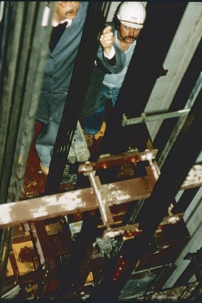 Zwei Arbeiter in einem Aufzugsschacht, wobei ein Mann mit weißem Schutzhelm und Schnurrbart deutlich zu sehen ist. Die Arbeitsplattform ist von Metallstrukturen und technischen Komponenten umgeben.