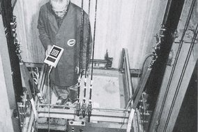 Schwarz-Weiß-Fotografie einer Person in Arbeitskleidung, die Steuerungsgeräte innerhalb eines Aufzugschachts oder mechanischen Systems bedient, wobei verschiedene Kabel und Strukturelemente sichtbar sind.