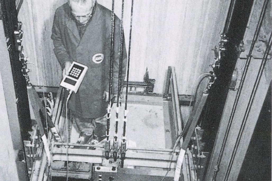 Schwarz-Weiß-Fotografie einer Person in Arbeitskleidung, die Steuerungsgeräte innerhalb eines Aufzugschachts oder mechanischen Systems bedient, wobei verschiedene Kabel und Strukturelemente sichtbar sind.
