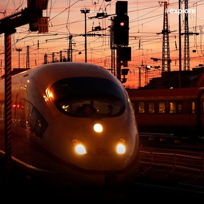 Weißer Hochgeschwindigkeitszug fährt bei Sonnenuntergang durch ein komplexes Netz von Oberleitungen und Signalmasten in dramatisch orangefarbenem Licht.