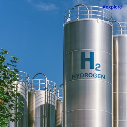 Mehrere große silberne Industrietanks mit "H₂ HYDROGEN" Beschriftung stehen unter blauem Himmel neben grünen Bäumen einer Wasserstoff-Speicheranlage.