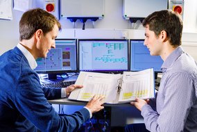 Zwei Techniker analysieren Daten auf Computerbildschirmen mit technischen Diagrammen, während sie eine Dokumentation in einem Ordner gemeinsam durchgehen.
