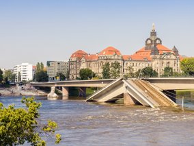 Eine teilweise eingestürzte Betonbrücke überspannt einen schlammigen Fluss, dahinter stehen historische Gebäude mit roten Dächern.