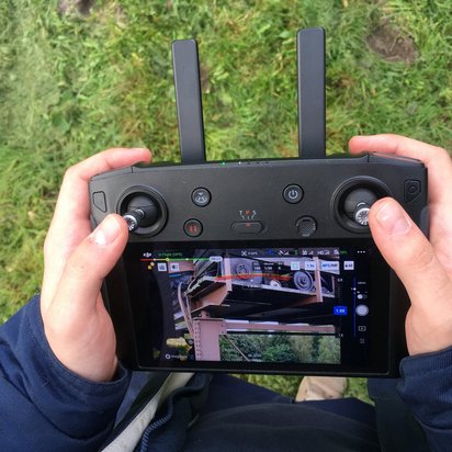 Zwei Hände halten eine schwarze DJI-Drohnen-Fernsteuerung mit Display, das eine Live-Kameraansicht zeigt. Im Hintergrund ist grünes Gras zu sehen, die Person trägt einen dunkelblauen Pullover.