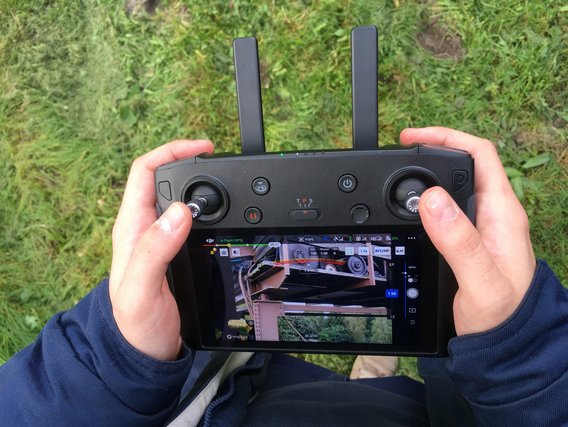 Zwei Hände halten eine schwarze DJI-Drohnen-Fernsteuerung mit Display, das eine Live-Kameraansicht zeigt. Im Hintergrund ist grünes Gras zu sehen, die Person trägt einen dunkelblauen Pullover.