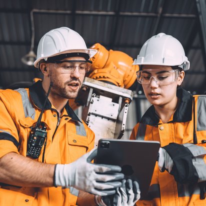 Zwei Fachkräfte in Warnkleidung und Schutzhelmen prüfen gemeinsam technische Daten auf einem Tablet in einer Industrieumgebung.