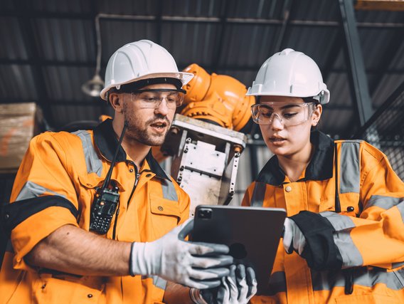 Zwei Fachkräfte in Warnkleidung und Schutzhelmen prüfen gemeinsam technische Daten auf einem Tablet in einer Industrieumgebung.