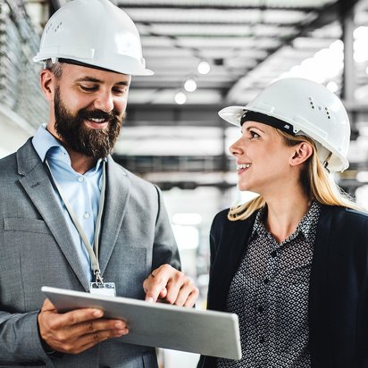 Zwei Personen mit Schutzhelmen stehen in einer Industriehalle und betrachten gemeinsam Informationen auf einem Tablet.