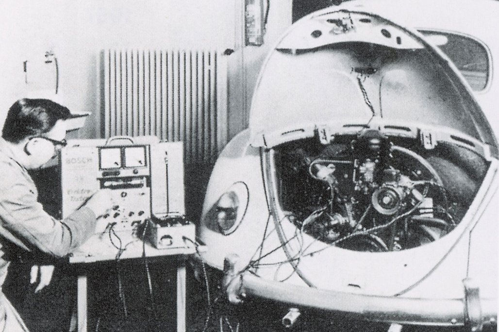  Schwarz-weiß Foto einer historischen Fahrzeugprüfung mit Messgeräten und einem VW Käfer in einer Werkstatt.